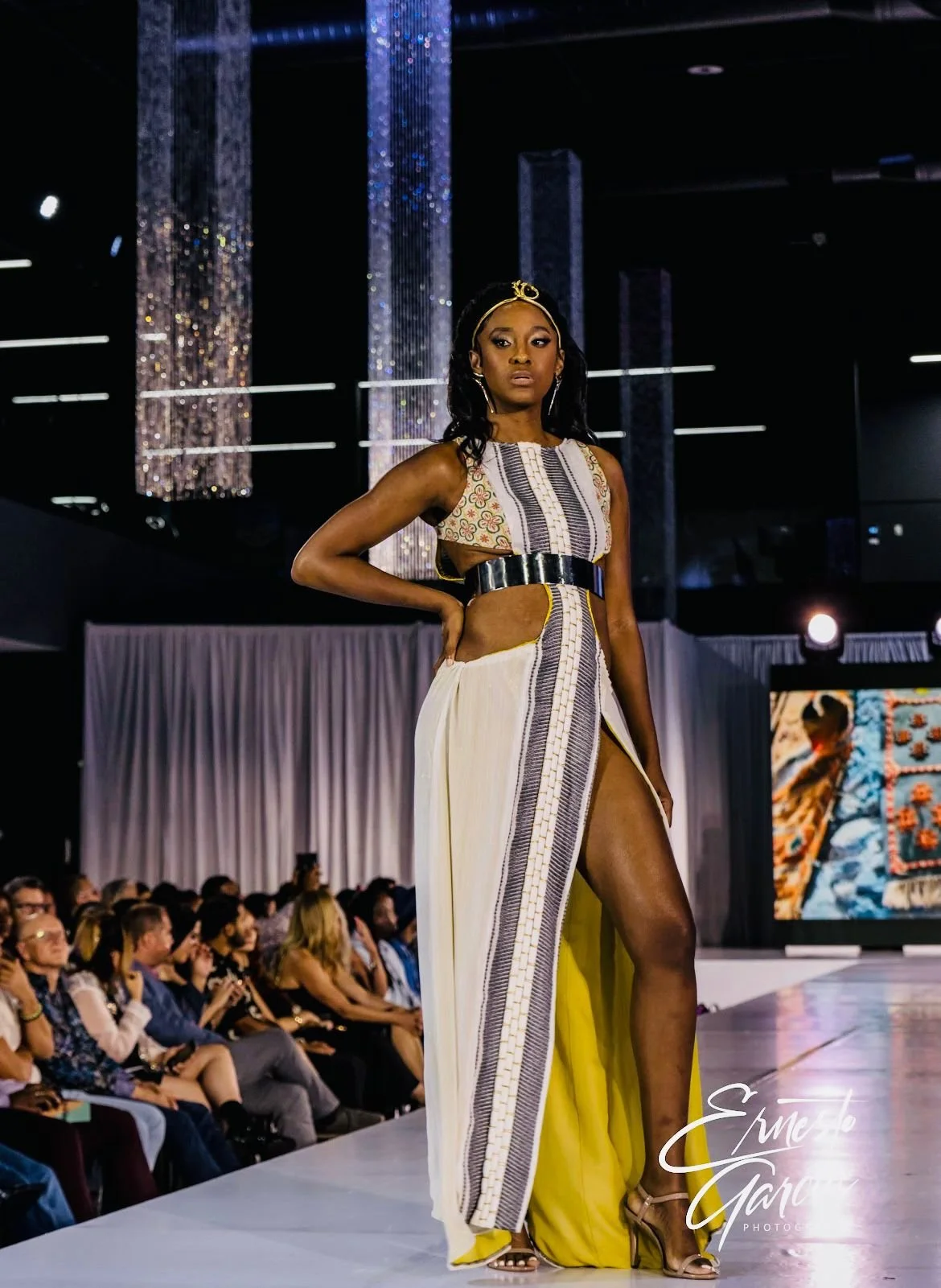 A female model walking on a runway during a fashion show, wearing a culturally inspired outfit with a slit and cutouts, with an audience in the background.