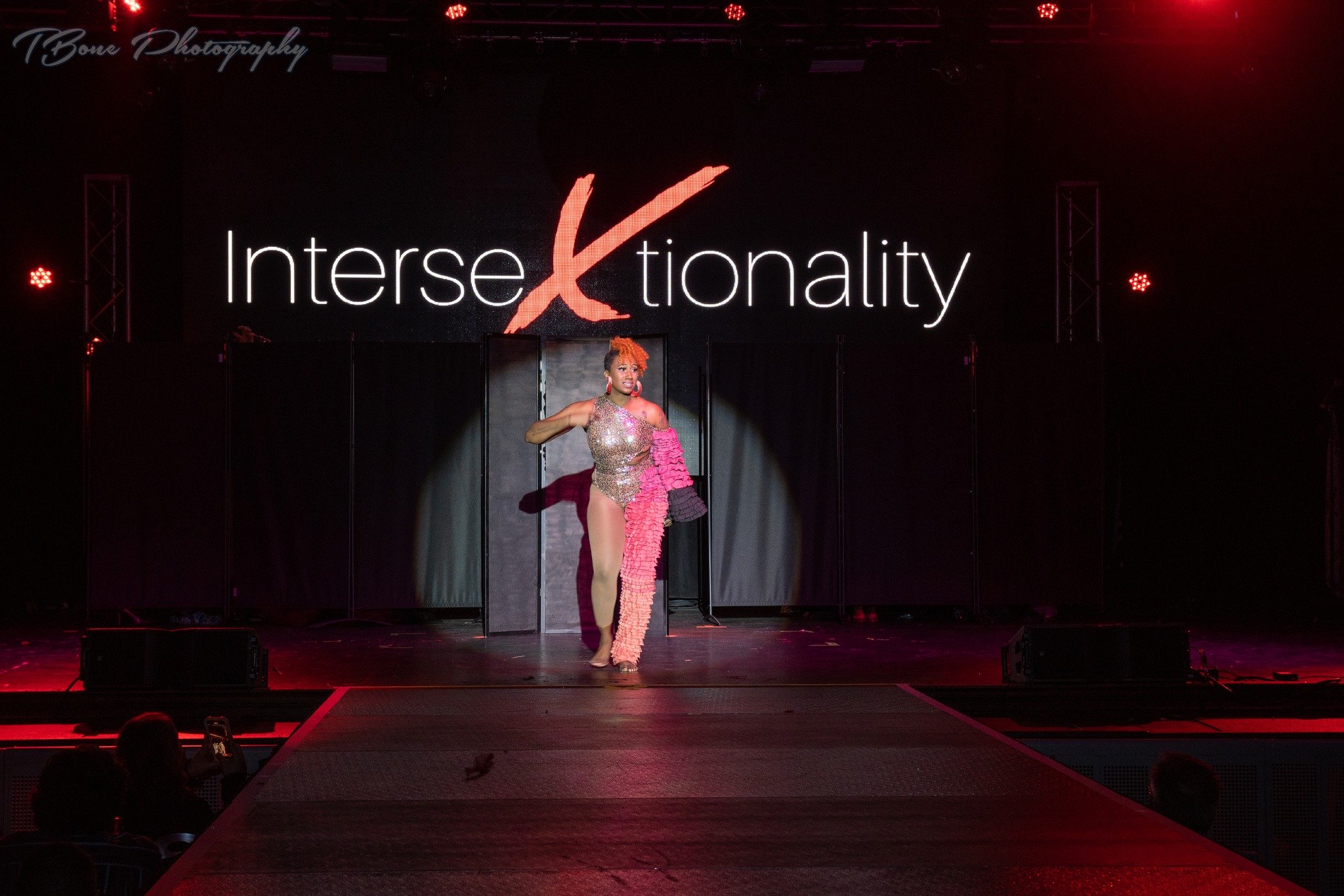 A performer on stage during a fashion show with a large screen displaying the word 'Intersectionality' in the background. The performer is wearing a sparkling outfit with a pink, ruffled accessory.