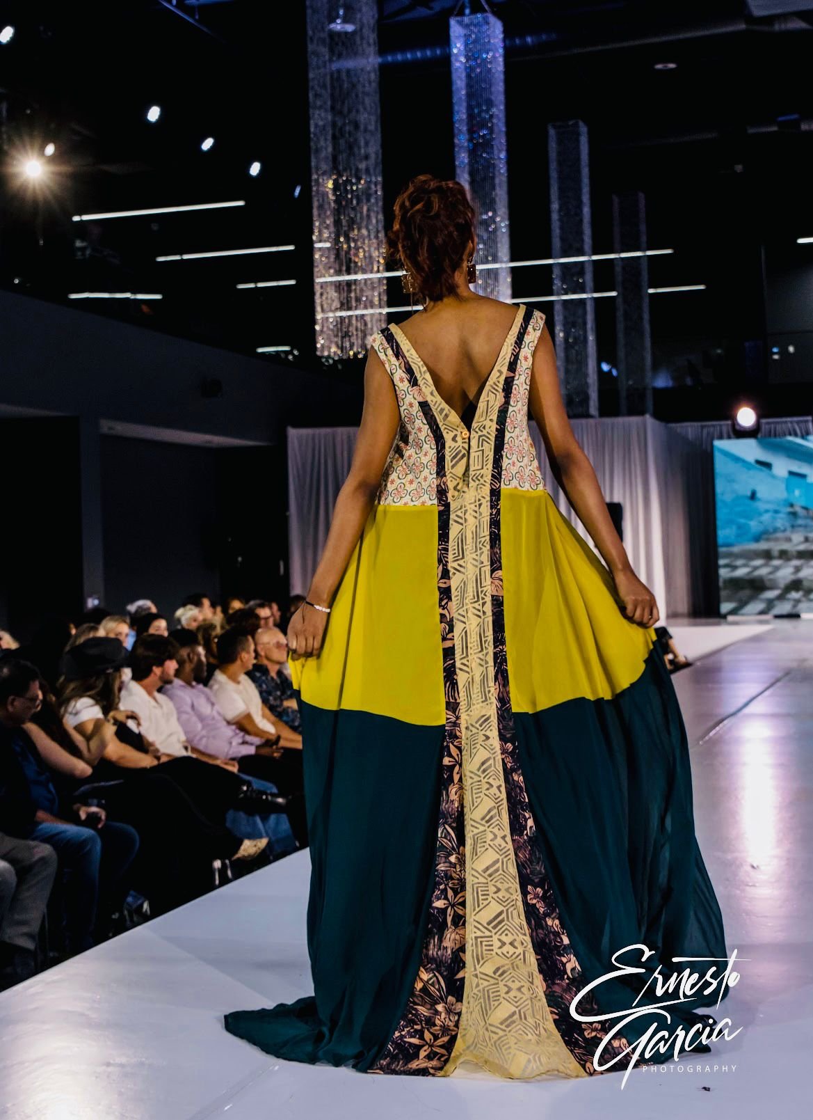 A model walks down a runway at a fashion show, wearing a colorful, floor-length dress with yellow, navy blue, and intricate patterned panels, in a large, dimly lit venue with an audience seated on either side of the runway.