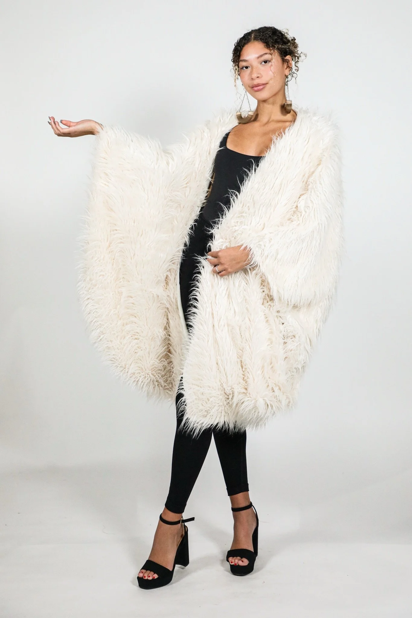 A young woman with curly hair wearing a fluffy white coat, black top, black leggings, and black high-heeled sandals, posing with one arm extended to the side against a plain white background.