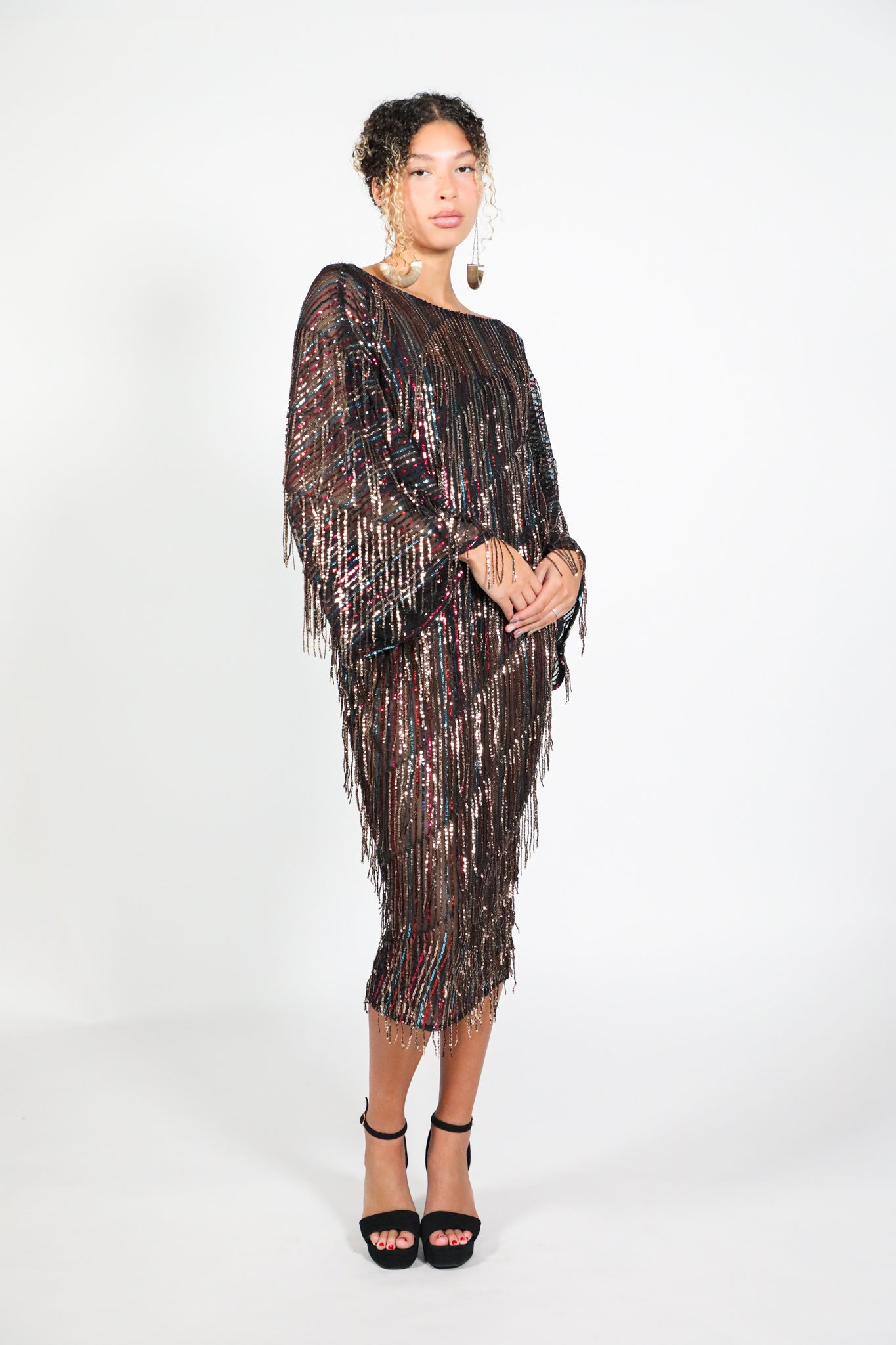 A woman wearing a sheer, black, sequined dress with long sleeves and a midi length, standing against a white background, paired with black high-heeled sandals and statement earrings.