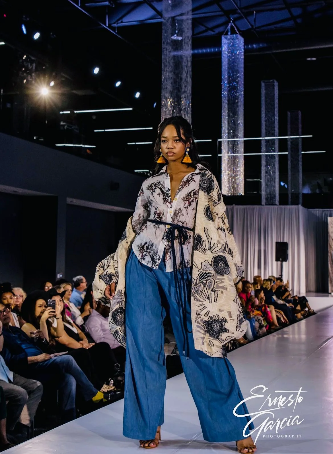 Fashion model walking on runway during fashion show, wearing patterned oversized shirt and blue wide-leg pants, audience seated watching.