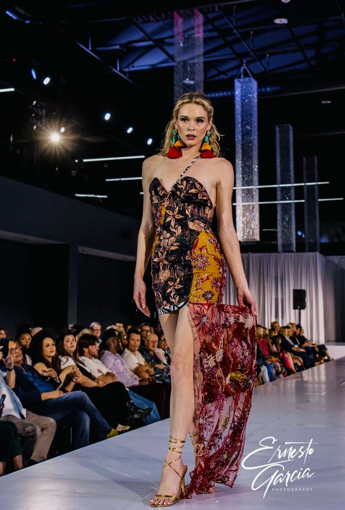 A female model walking on a runway during a fashion show, wearing a colorful dress with floral and embroidered details, large colorful earrings, and gold high-heeled sandals, with an audience watching in the background.