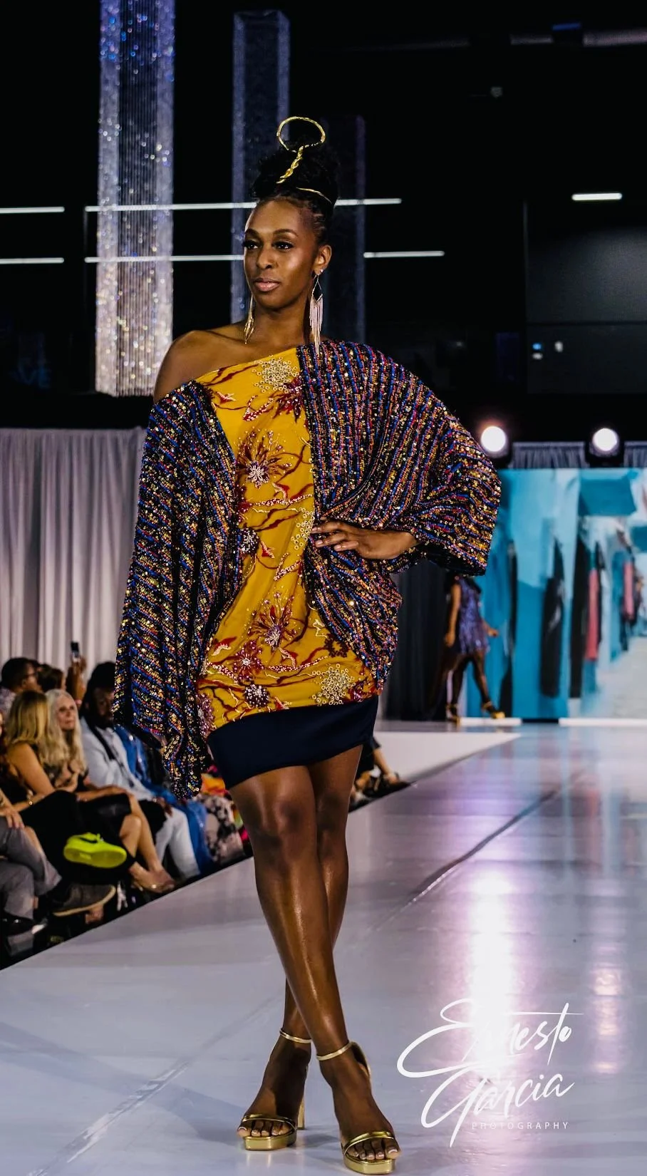 Model walking down runway in African-inspired yellow and multicolored beaded outfit with patterned shawl, in a fashion show setting.