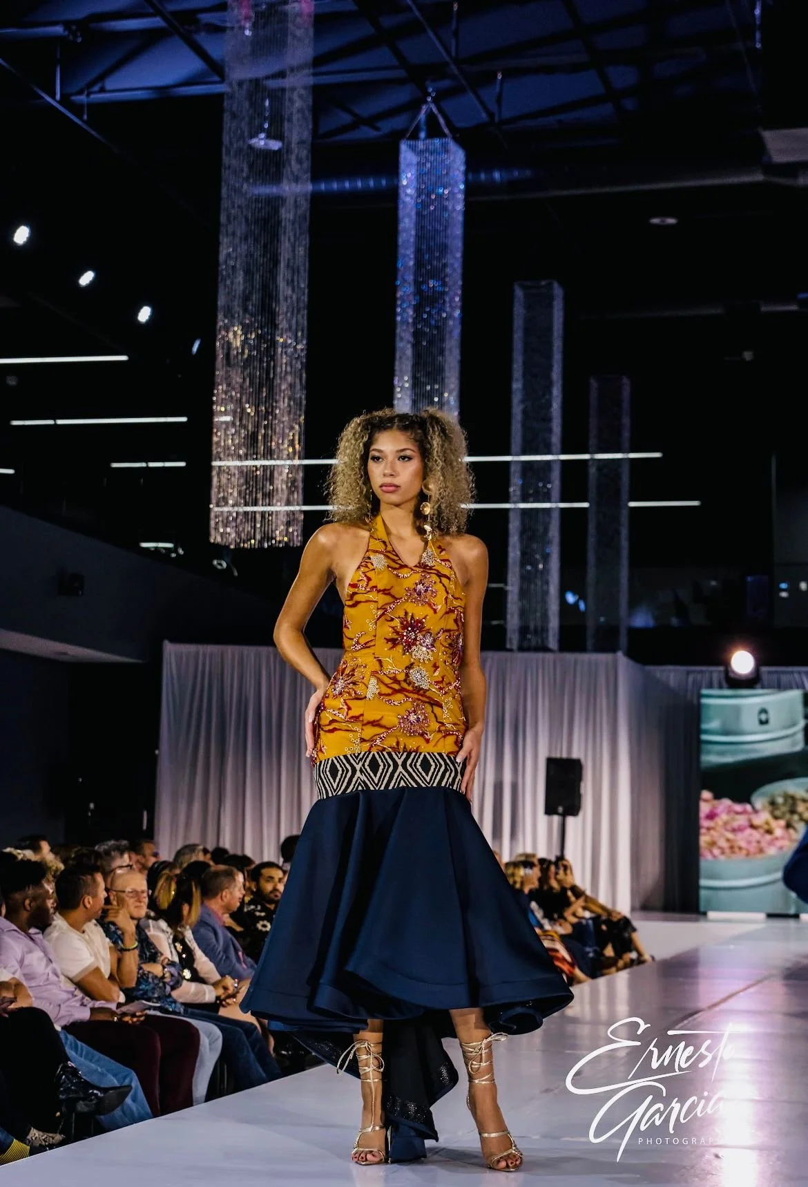 A model walking runway at a fashion show, wearing a colorful top with a dark blue ruffled skirt, with audience watching in the background and chandeliers hanging from the ceiling.