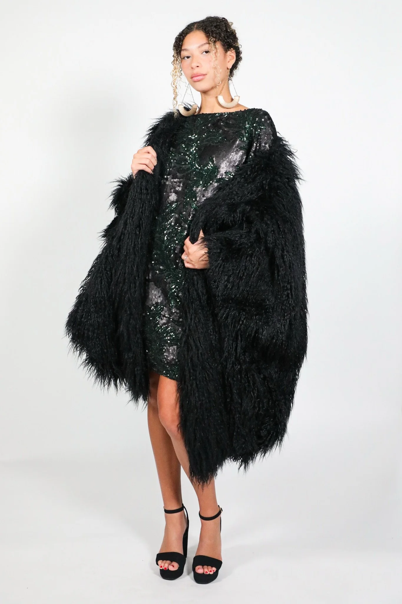 A woman in a black sequin dress and black high heels, wearing large earrings and a black fur coat, poses against a plain white background.