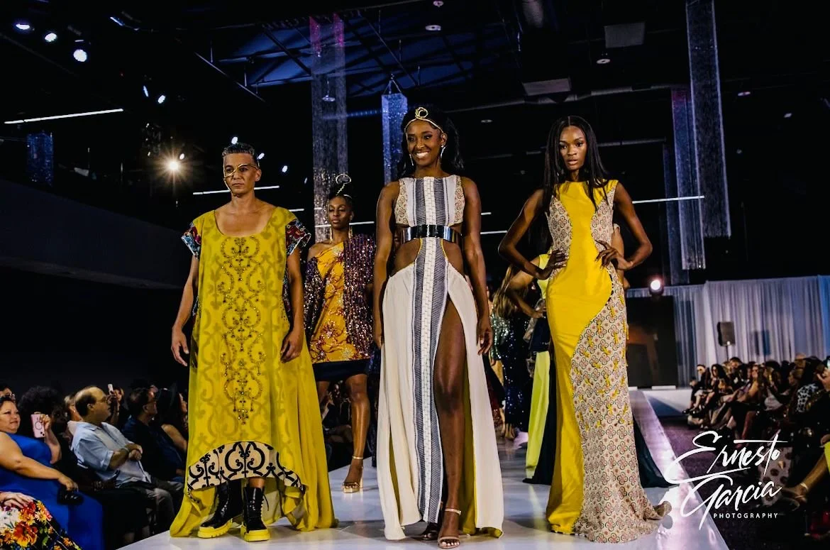 Models walking down the runway during a fashion show, wearing vibrant and patterned dresses, with audience watching.