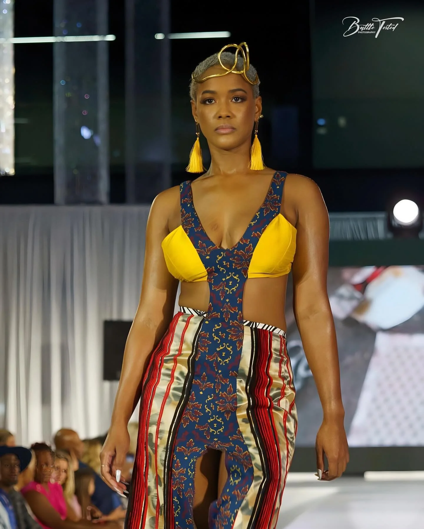 A woman walking on a fashion runway wearing a colorful textile-patterned outfit with yellow and blue accents, large yellow tassel earrings, and yellow sunglasses on her head.