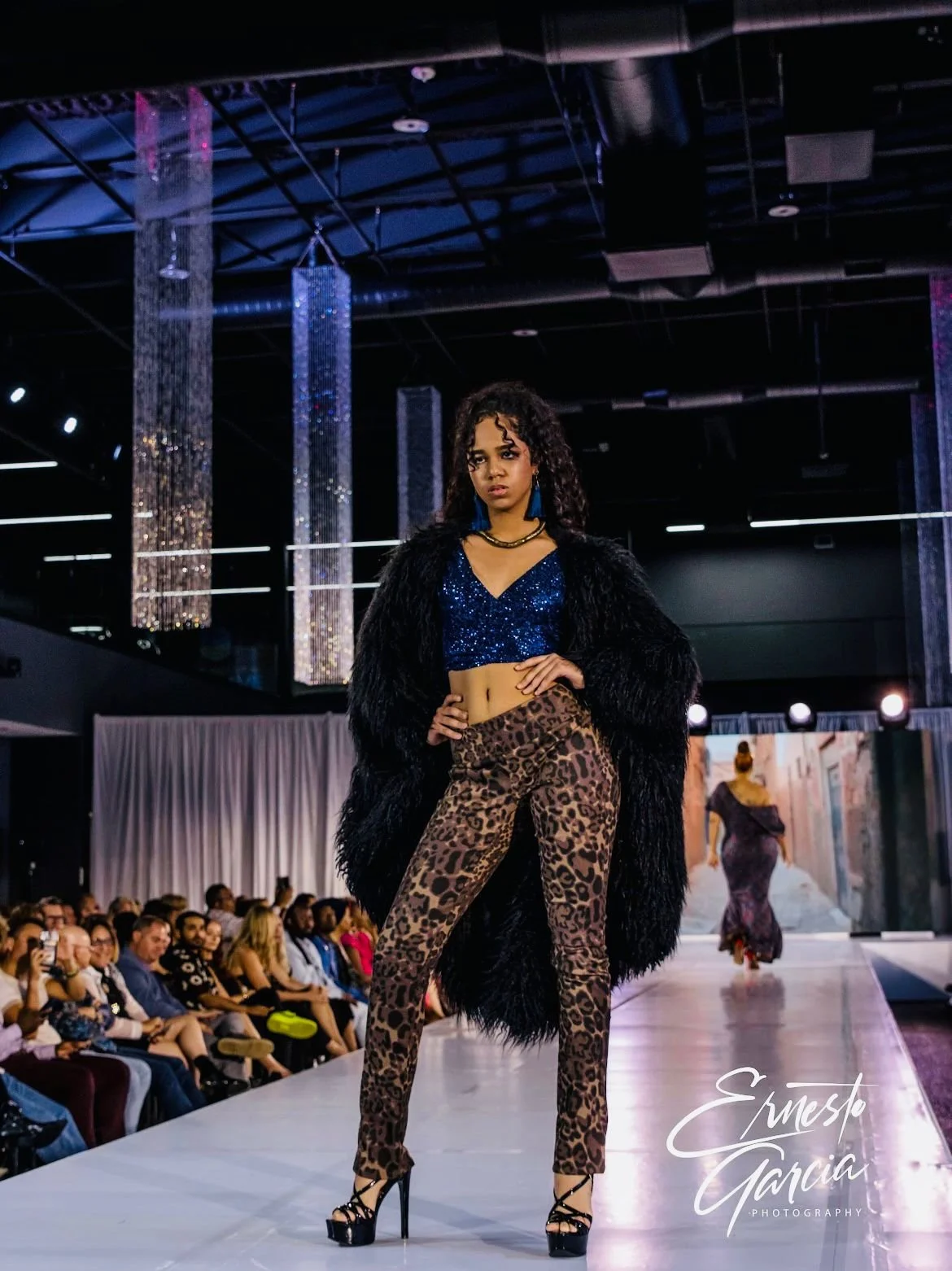 Fashion model walking on runway at a fashion show wearing leopard print pants, blue glittery crop top, and black fur coat, audience seated along the runway, stage with hanging lights and a large screen in the background