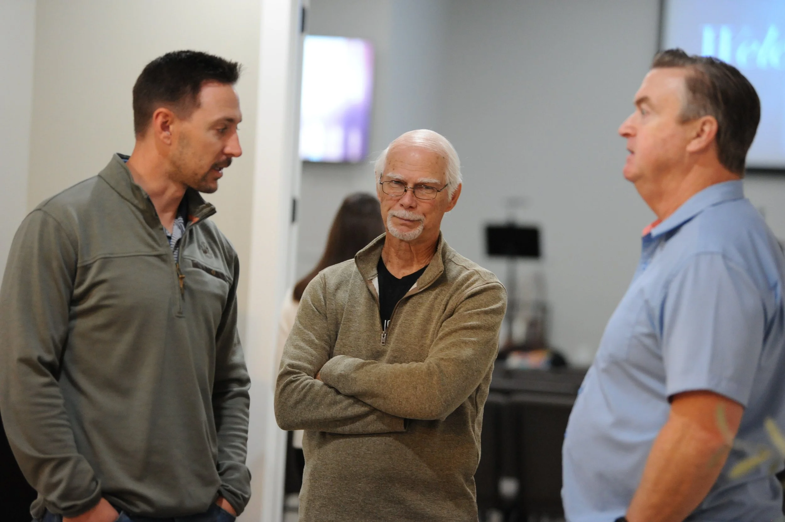 Three men having a conversation in a room with a monitor and people in the background.