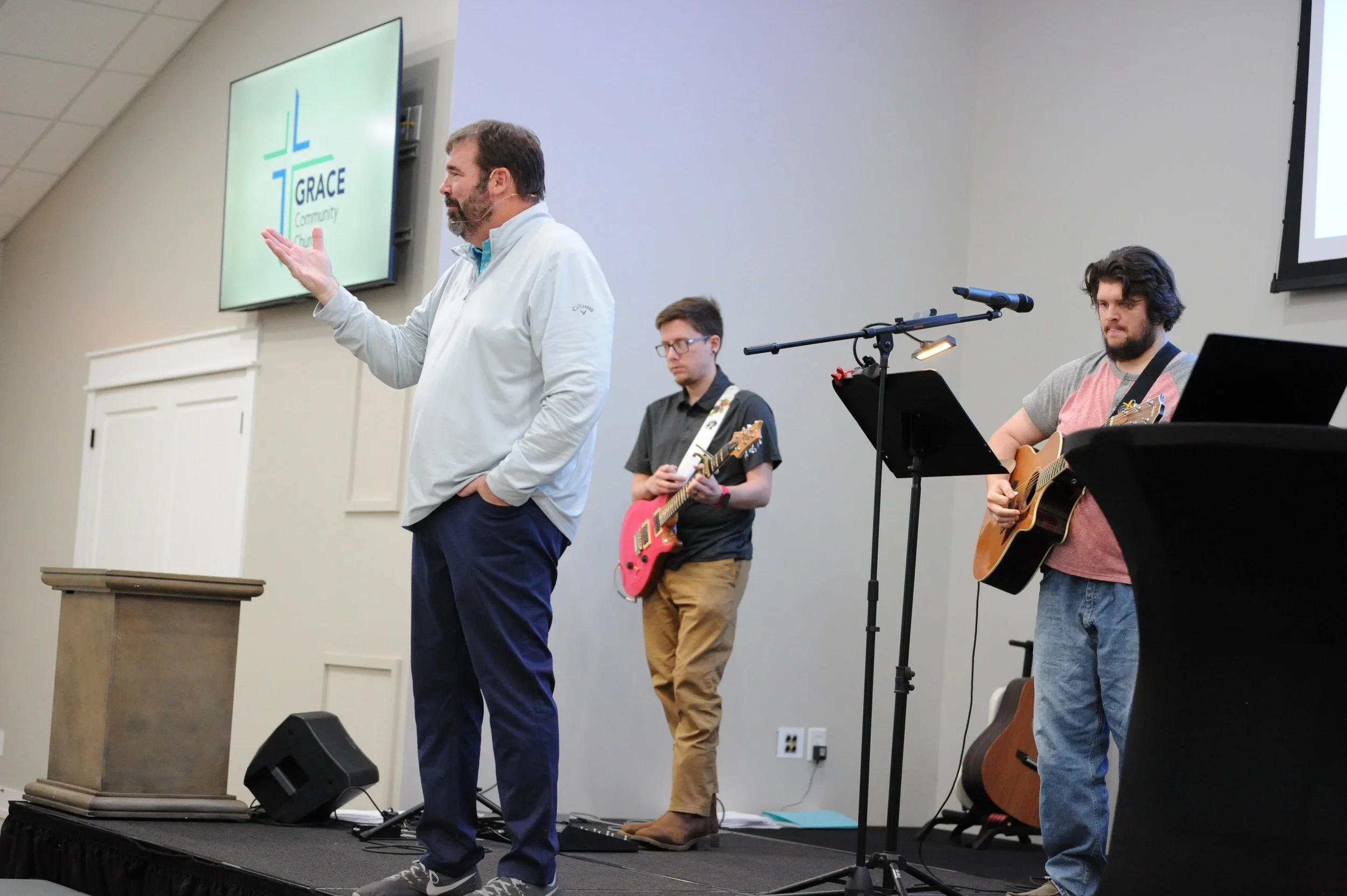 A man speaking at a church event with two musicians playing guitars beside him. The church logo, 'GRACE Community Church,' is displayed on a large screen behind them.