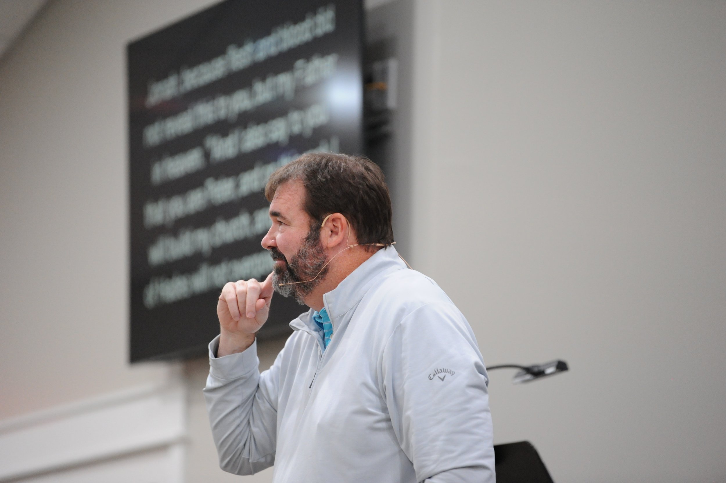 Man with brown hair and beard wearing a white jacket, standing in front of a blackboard, in profile view, speaking or presenting.
