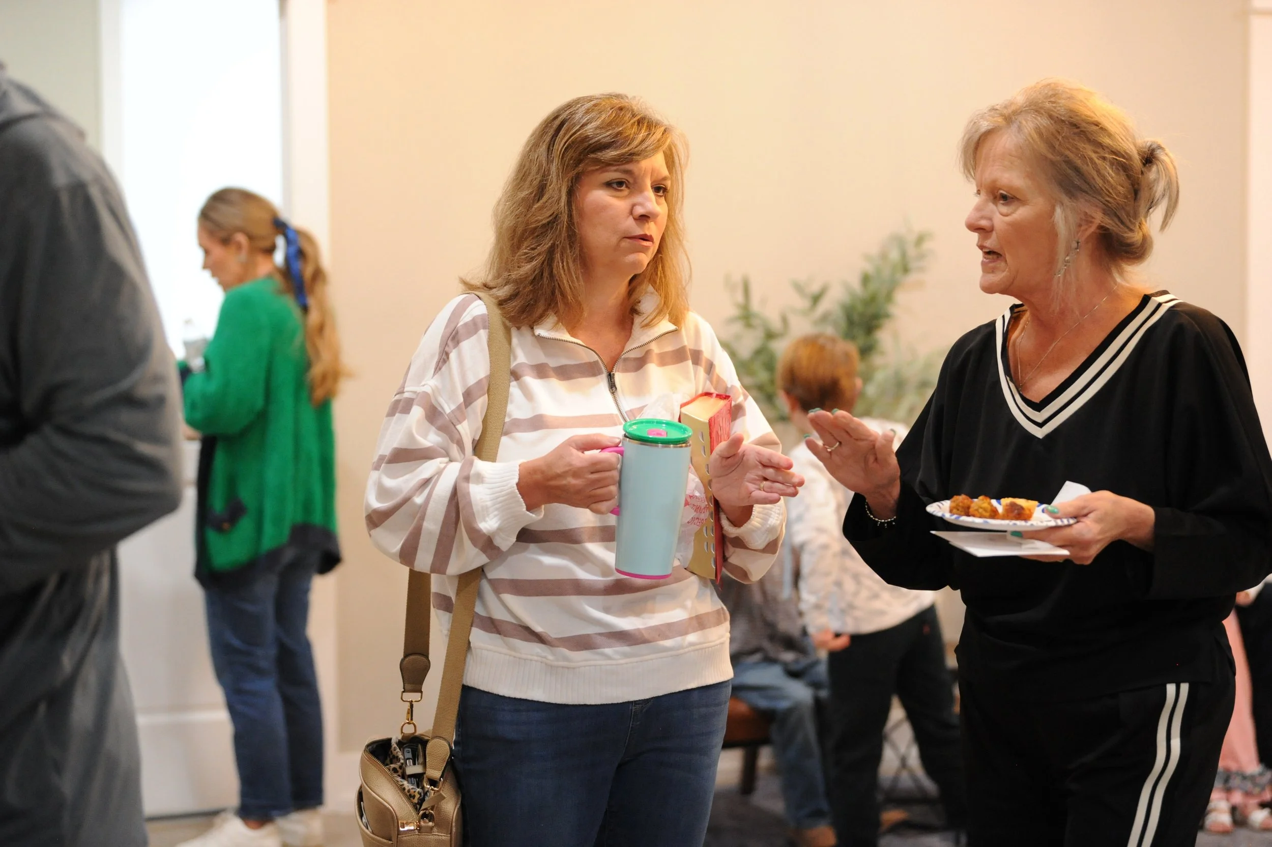Two women talking at a social gathering, one holding a plate with food, the other holding a mug and a book.