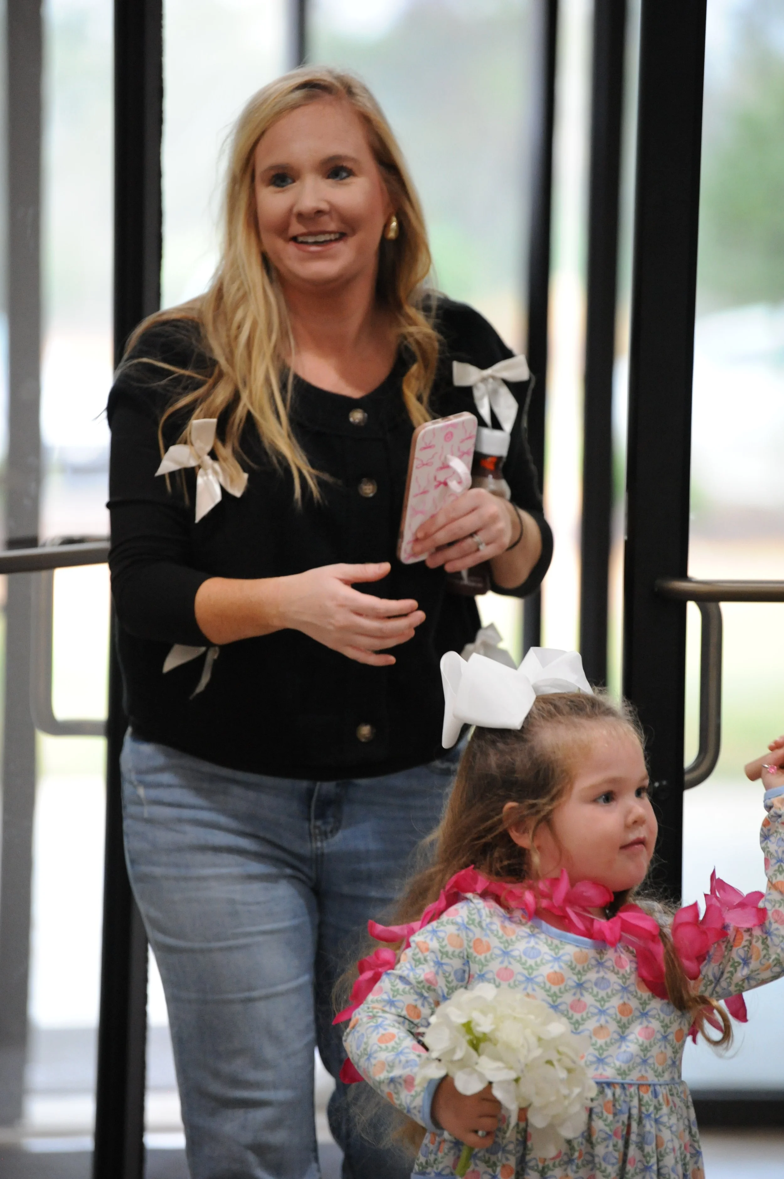 A woman with long blonde hair and a black jacket with white bows, smiling and holding a phone and a drink, standing behind a young girl with curly hair, a white bow, and a floral dress, holding a bouquet of flowers and wearing a pink lei, inside near glass doors.