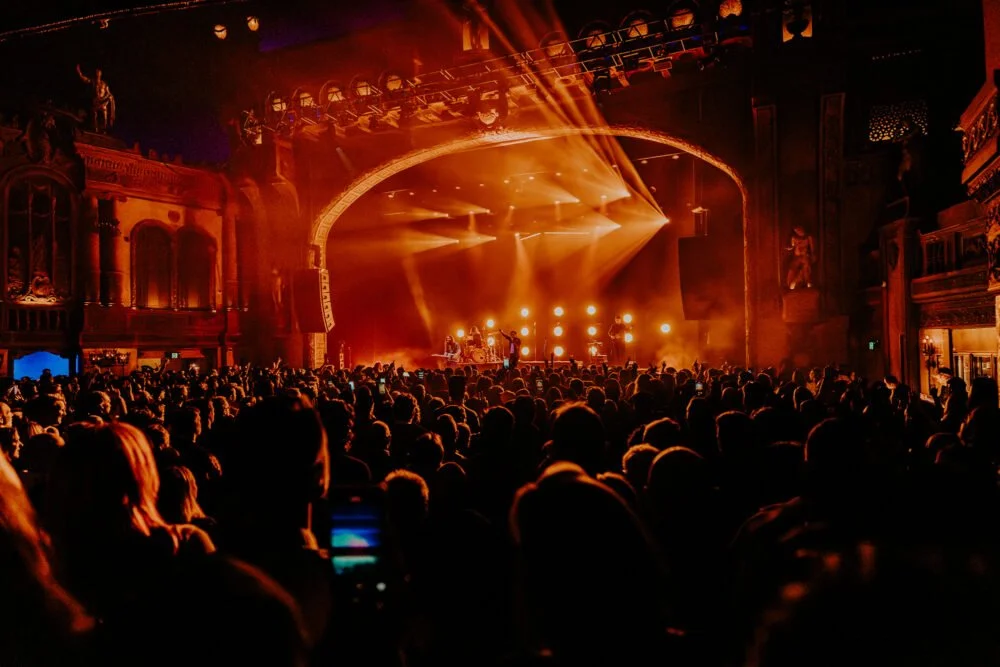 A crowded concert hall with a stage illuminated by bright orange and yellow lights, performing musicians, and an audience watching the show.