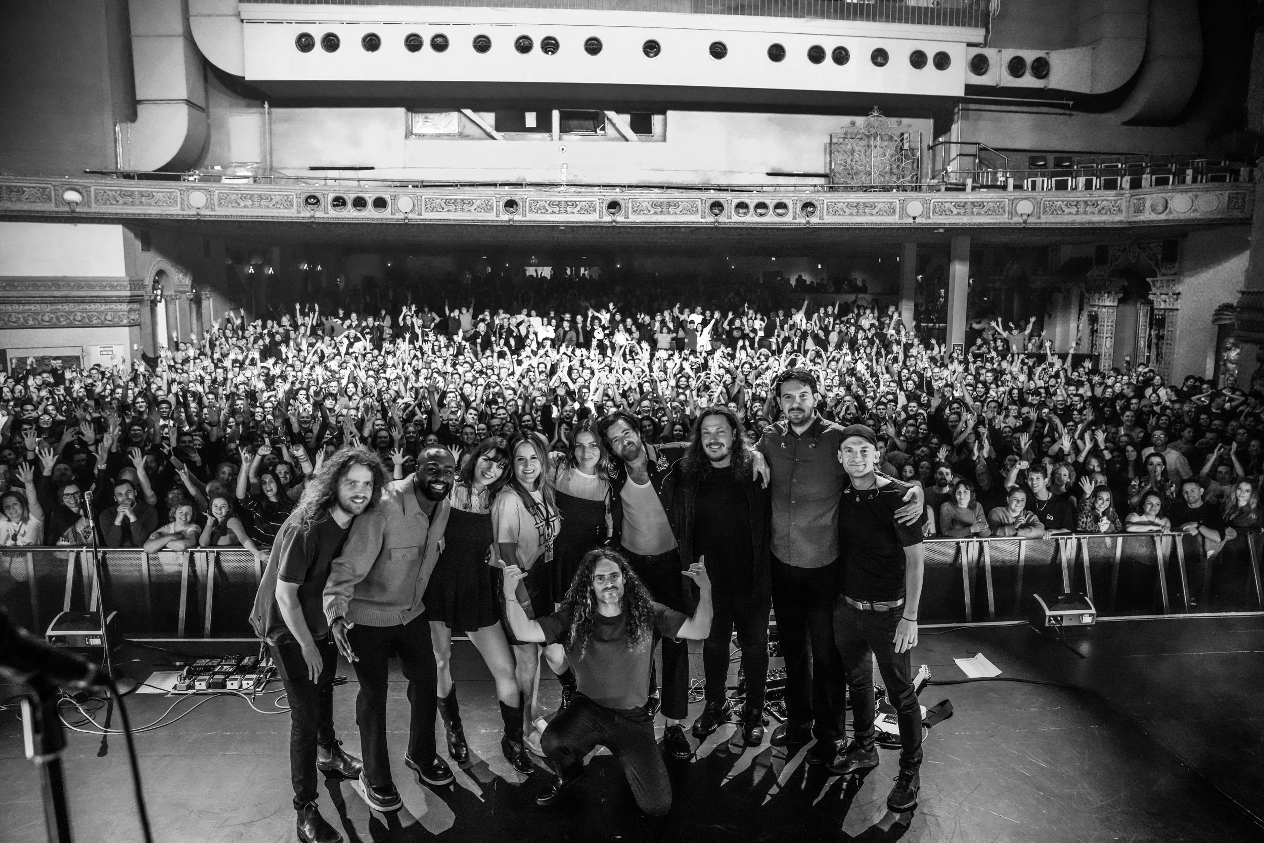 Group of nine people (staff and Holy Holy band members) posing on stage in front of a large audience inside a theater, black and white photo.