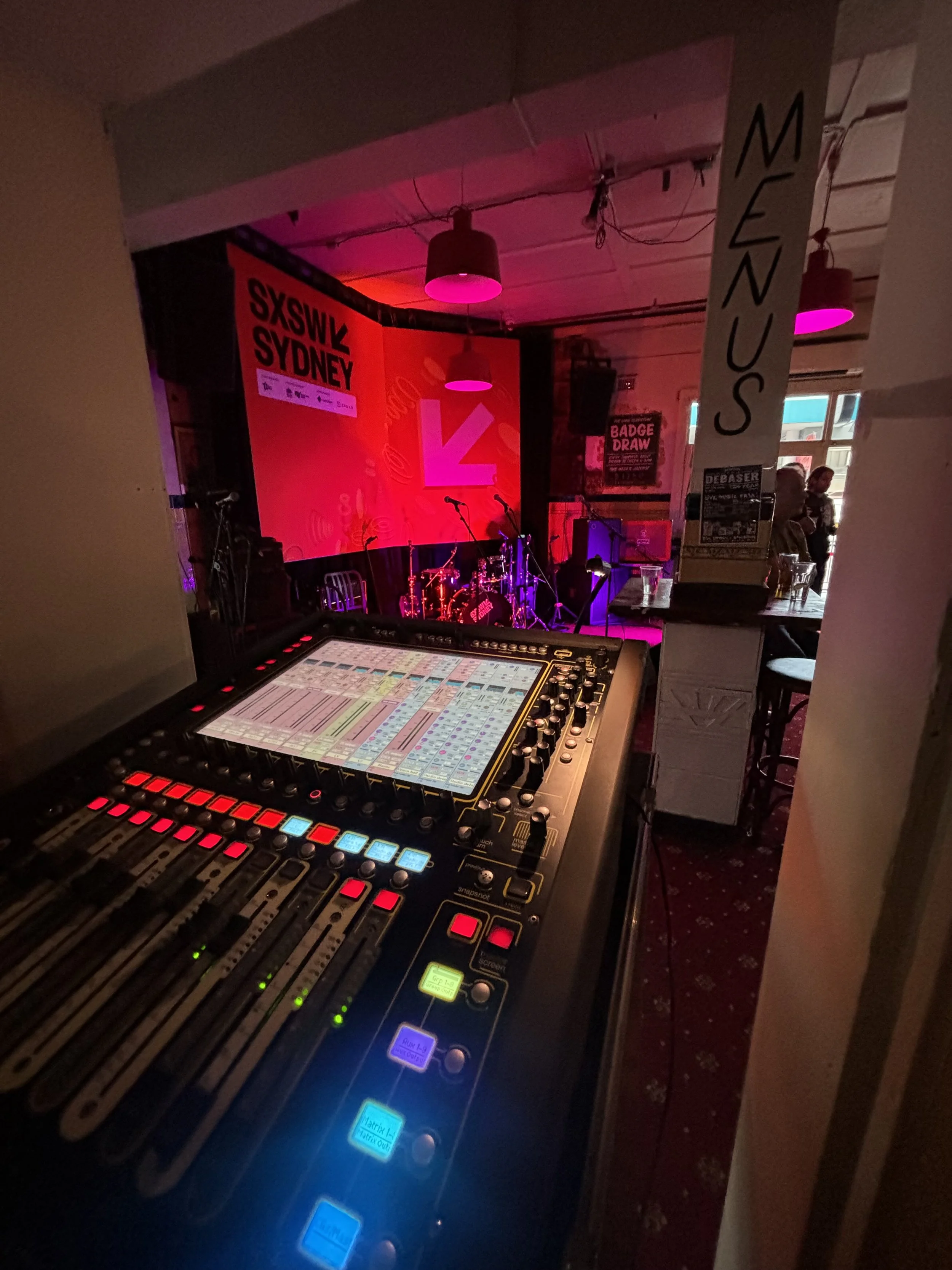View of a sound mixing console in the foreground, with a stage featuring drums and microphones in the background at SXSW Sydney event with pink and purple lighting.
