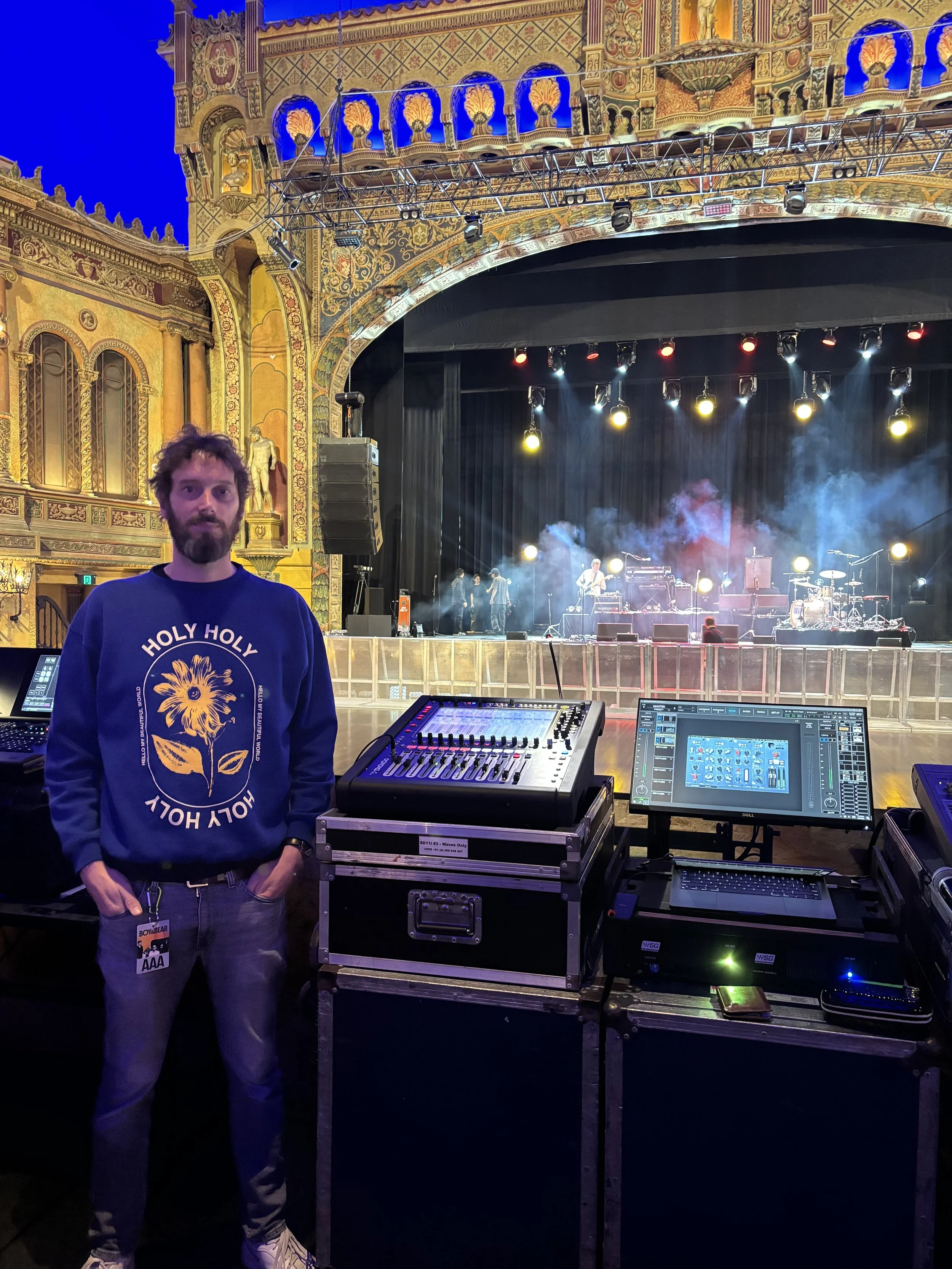 Photo of Adam Hignett. Man standing next to sound equipment in a concert hall with an ornate stage, stage lights, and musicians performing on stage.