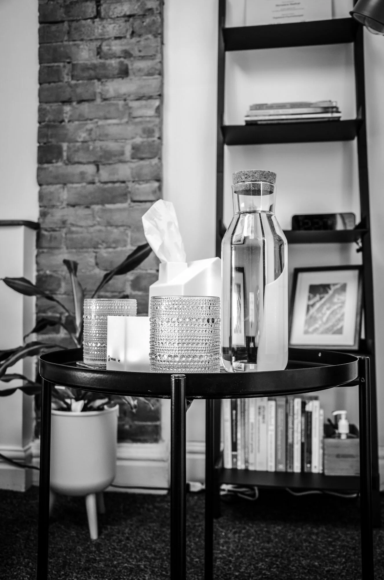 A round black table with a glass pitcher of water, two textured glasses, a tissue box, and a container of tissues. Background includes a brick wall, a bookshelf, a potted plant, framed pictures, and a digital clock.
