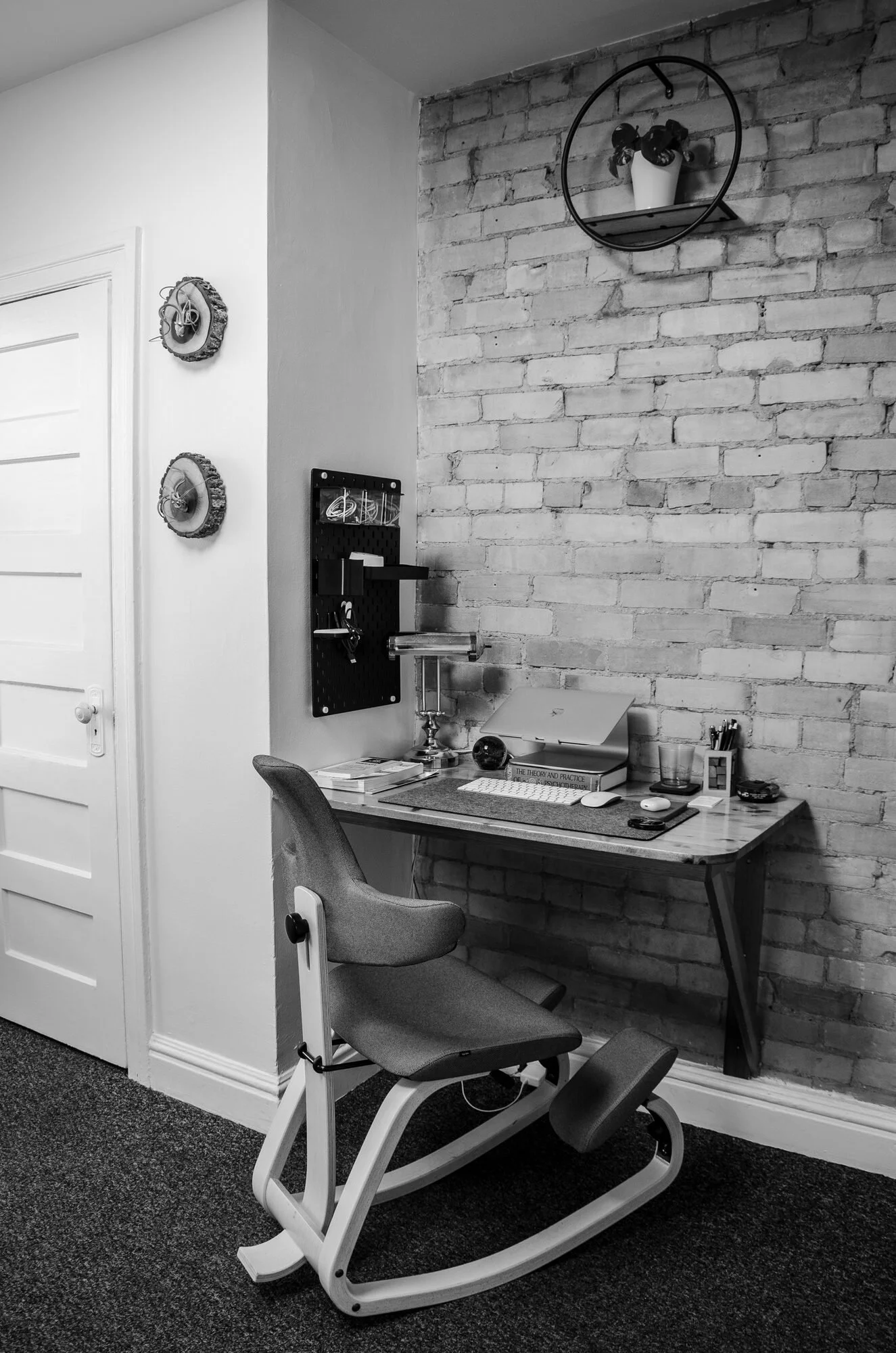 An office corner with a brick wall, a wooden desk, an office chair, and decorative wall items, including a round wall shelf with a potted plant and two wall-mounted hooks.