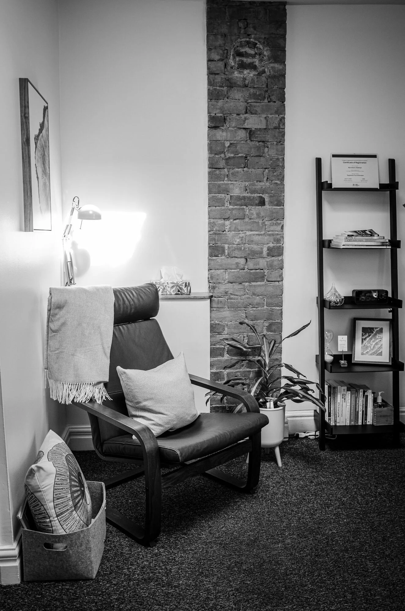 A cozy corner in a room with a black leather armchair, a cushion, a throw blanket, a potted plant, a bookshelf with books and decorative items, a framed certificate, and a brick wall, all in black and white.