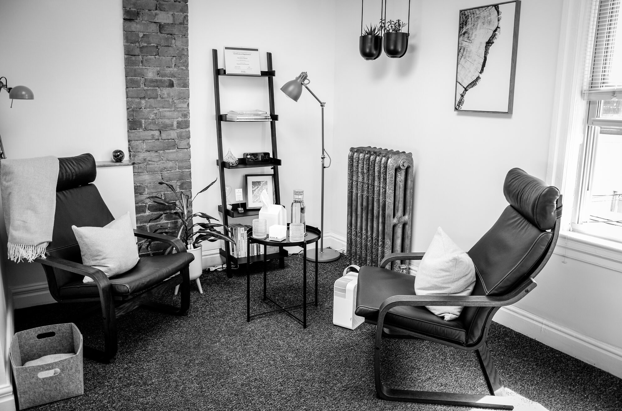 A cozy living room with two black leather chairs, a small round table with candles, a bookshelf, a plant, and wall art. There are windows with blinds, a radiator, and decorative hanging planters.