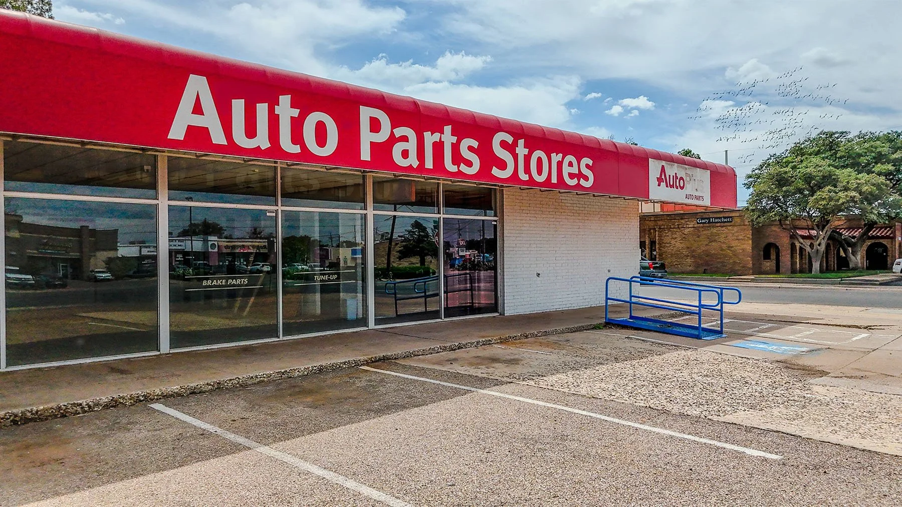 Front of an auto parts store with large red sign reading 'Auto Parts Stores' and glass windows, with parking spaces in front and a blue shopping cart frame to the side.