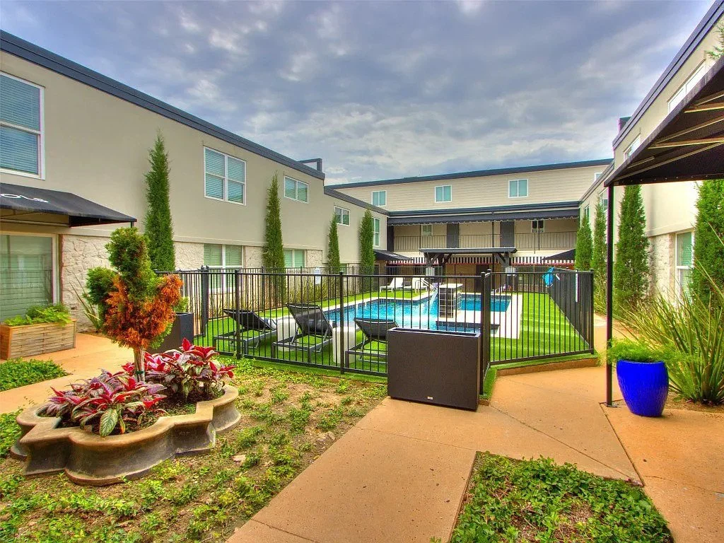 Residential apartment complex with a fenced swimming pool, lounge chairs, and landscaped yard with plants and trees.