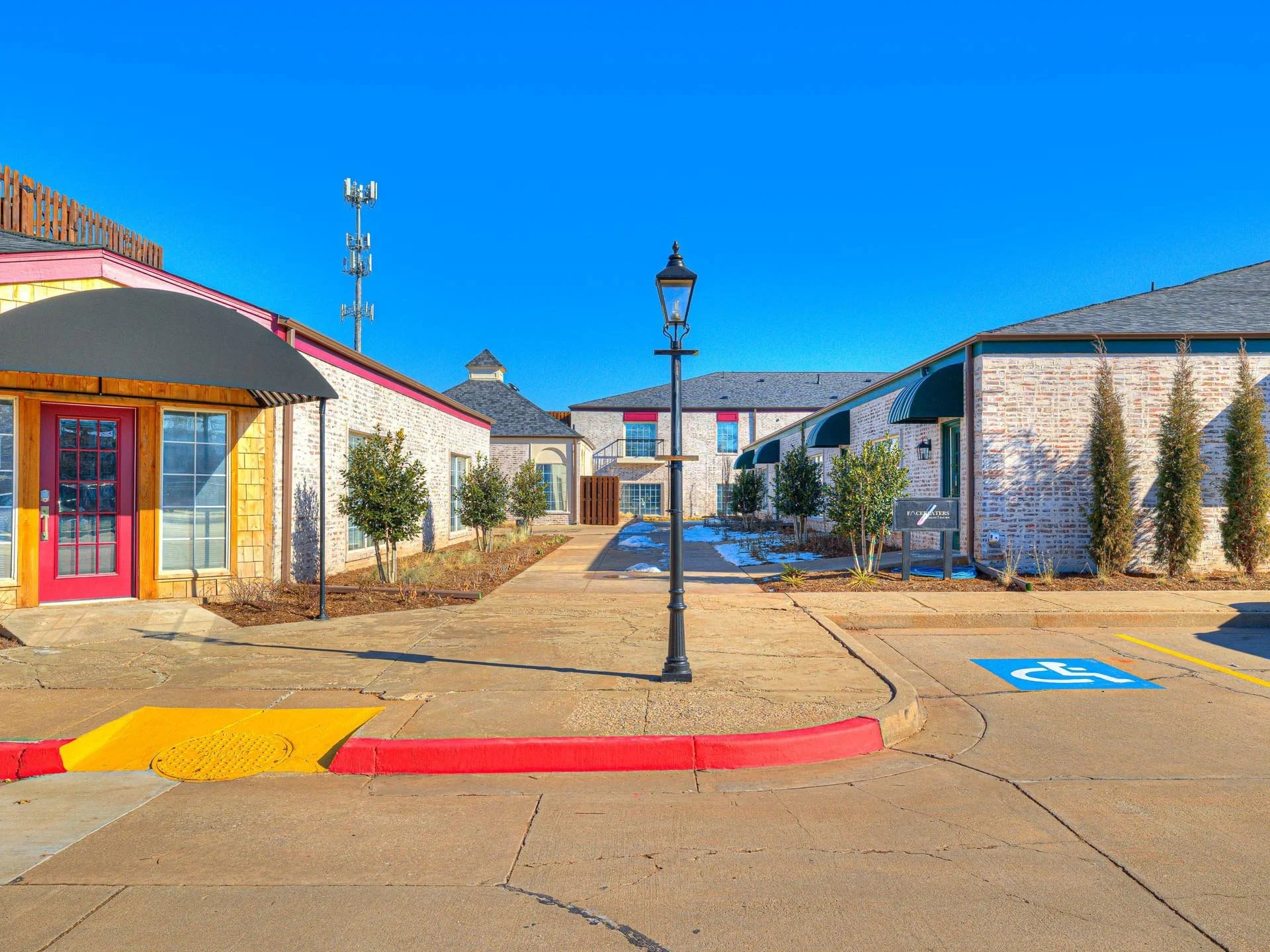 Exterior view of a senior apartment complex with a sidewalk, handicap parking space, and landscaped area with trees and shrubs. Clear blue sky in background.