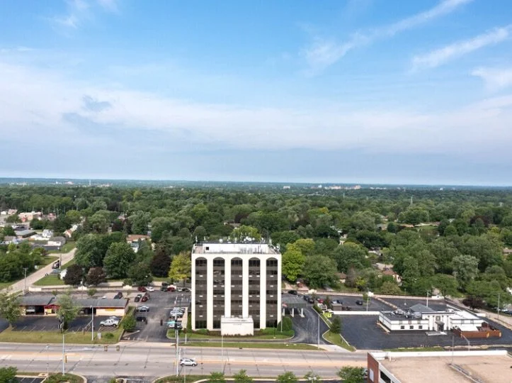 Aerial view of a cityscape with a large white multi-story building surrounded by parking lots and trees, under a partly cloudy sky.