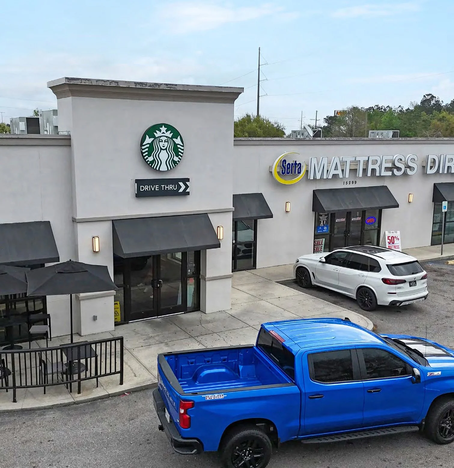 Exterior view of a retail store with a Starbucks sign, a Serta mattress store, outdoor seating, and parked cars including a blue pickup truck and a white SUV.