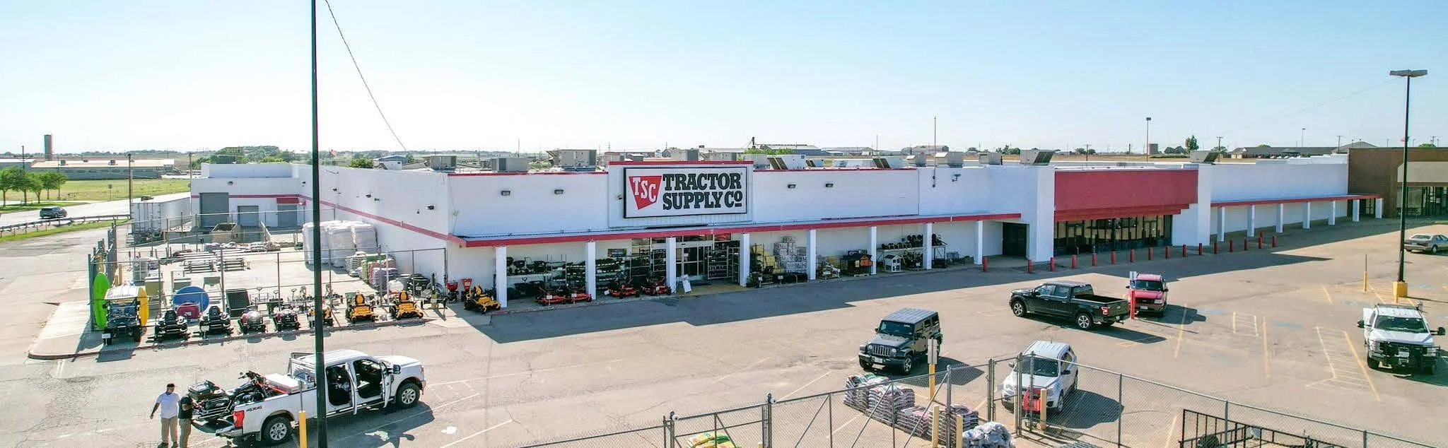 Exterior of Tractor Supply Co. store with parked vehicles, outdoor equipment, and a fencing section, under clear sky.