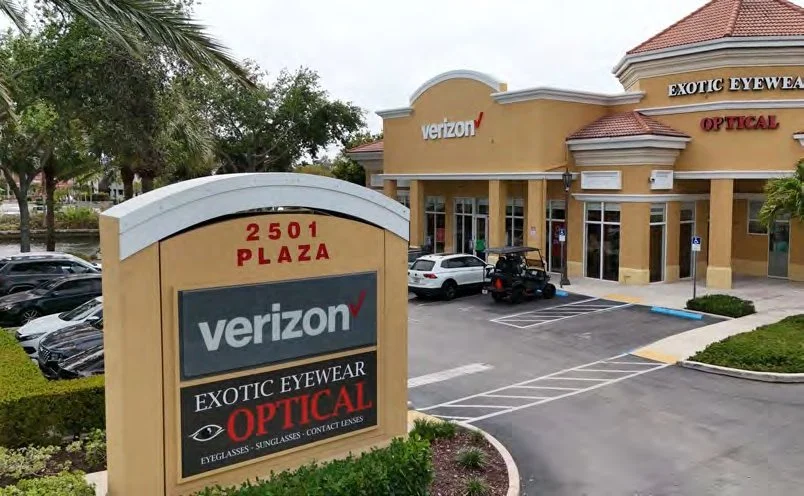 Strip mall with Verizon store and an optical eyewear shop, parking lot with cars, palm trees, and a beige building with a red roof.