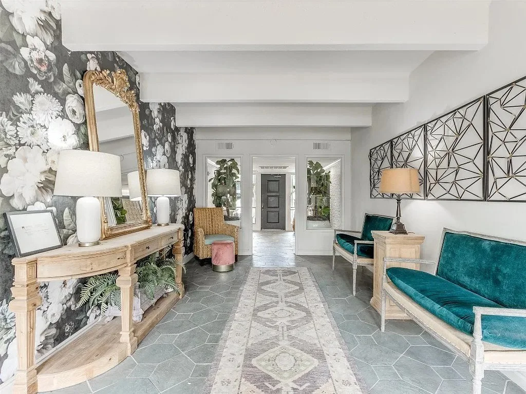 Bright, modern entryway with floral wallpaper on the left, a large gilded mirror, and a console table with lamps. Blue velvet seating on the right and a patterned runner rug along the tiled floor. Plants and decorative wall art complete the space.