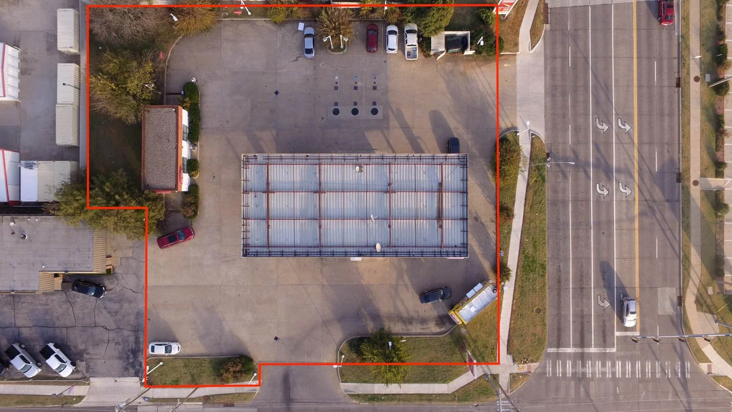 aerial view of a parking lot with various vehicles, a small building, and a large metal structure, surrounded by streets and sidewalks with some trees and grass, with boundary marked in red