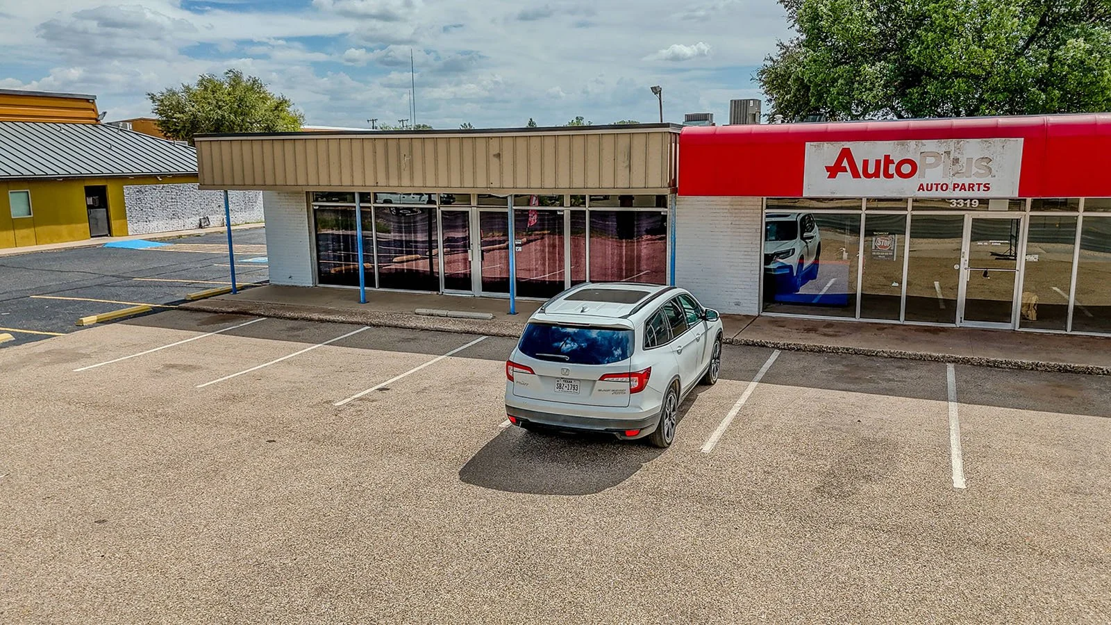 Empty parking lot in front of an AutoPlus auto parts store with a white SUV parked in front.