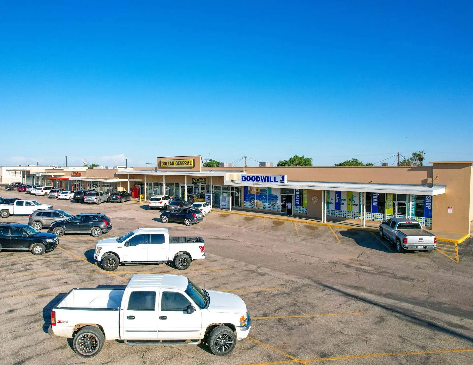 Parking lot in front of a small shopping plaza with stores like Dollar General and Goodwill, scattered cars parked on a clear day with blue sky.