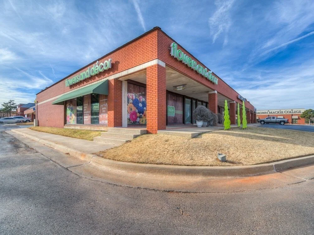 A brick storefront with a sign that reads "flowers and decor" and large windows displaying floral arrangements. In front of the store, there are green decorative trees and an open parking lot under a blue sky.