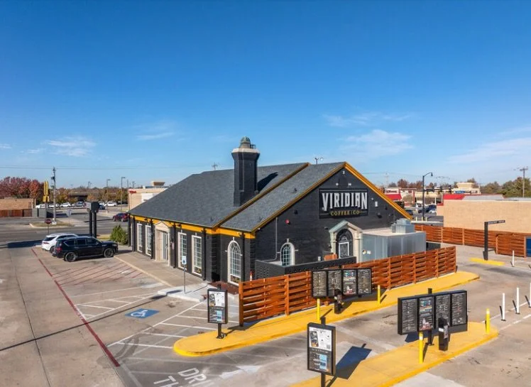 A coffee shop named 'Viridian Coffee Co.' in a standalone building with a black exterior, large windows, and a gray roof. The parking lot in front has several parking spaces, some occupied, and a few digital menu boards near the entrance. There is a