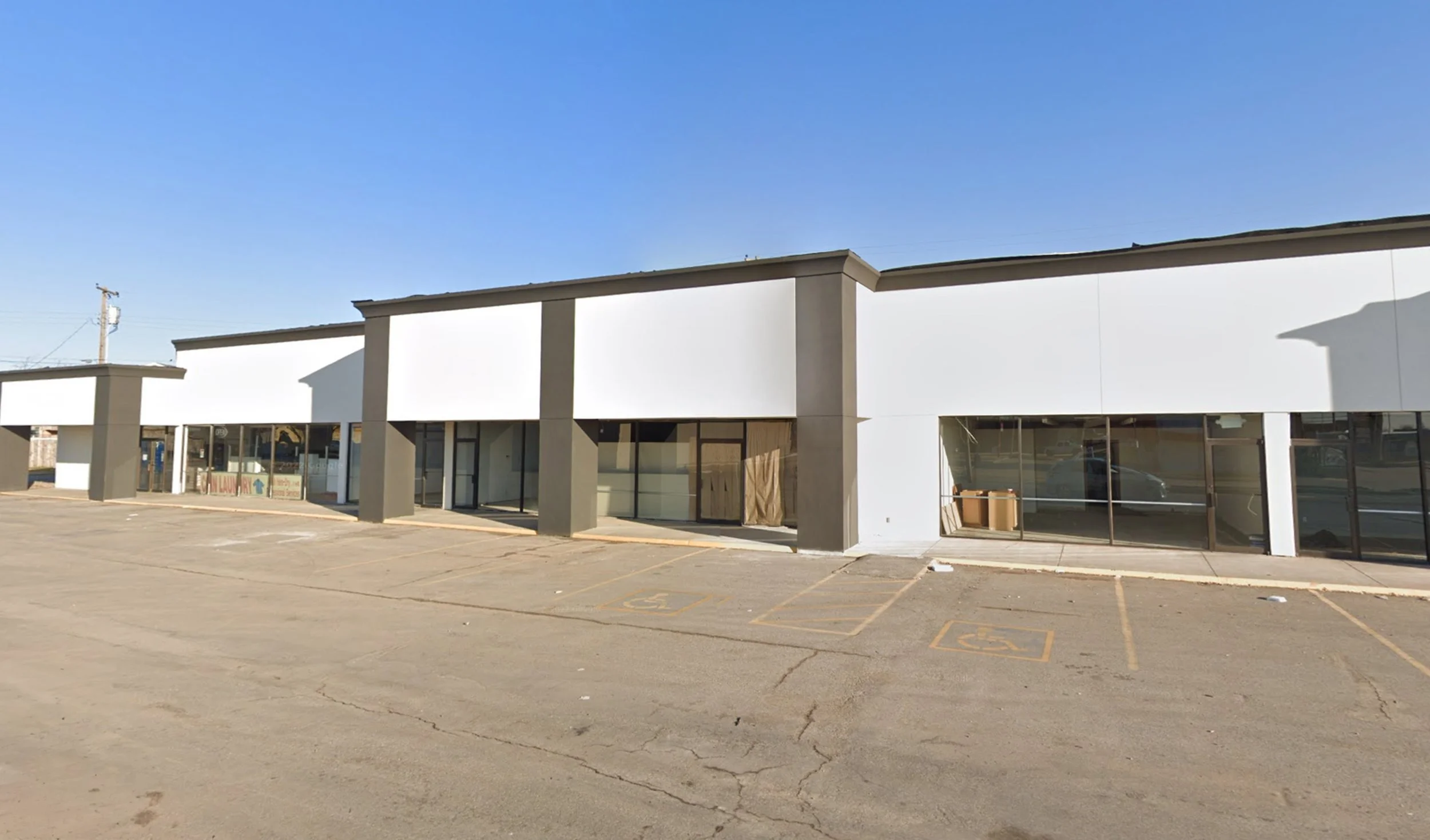 Empty parking lot in front of a row of empty storefronts with large glass windows and brown doors, under a clear blue sky.