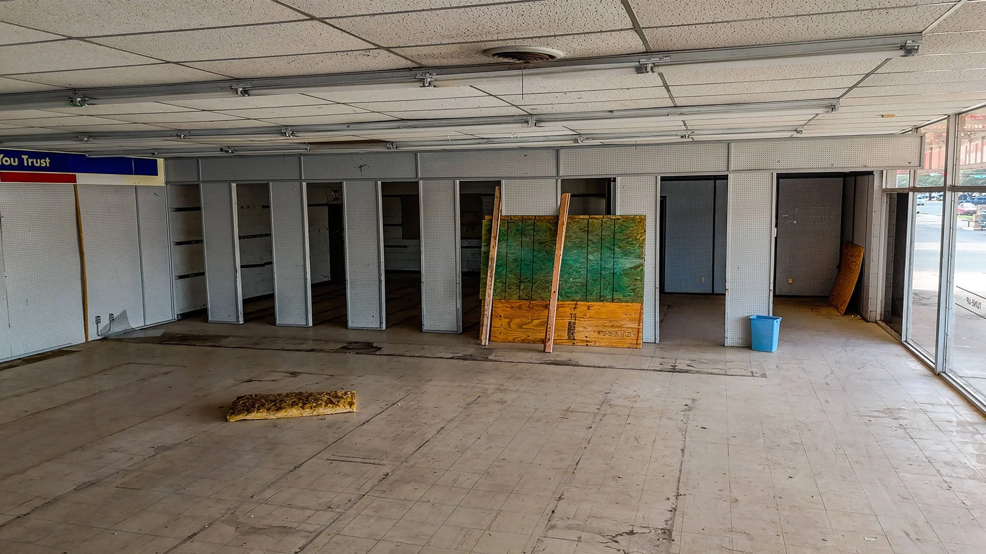 An empty storefront with bare walls and flooring, debris on the ground, and a few wooden and painted boards leaning against the wall. A blue trash can and an open entrance with glass doors are visible.
