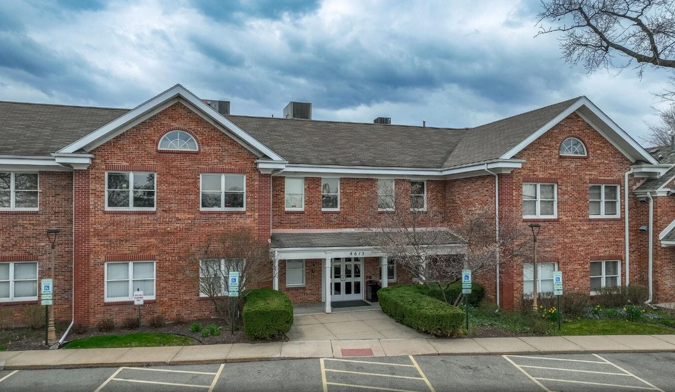 A three-story brick residential building with multiple windows and a main entrance with glass doors, surrounded by a sidewalk, parking spaces, and some trees and bushes.