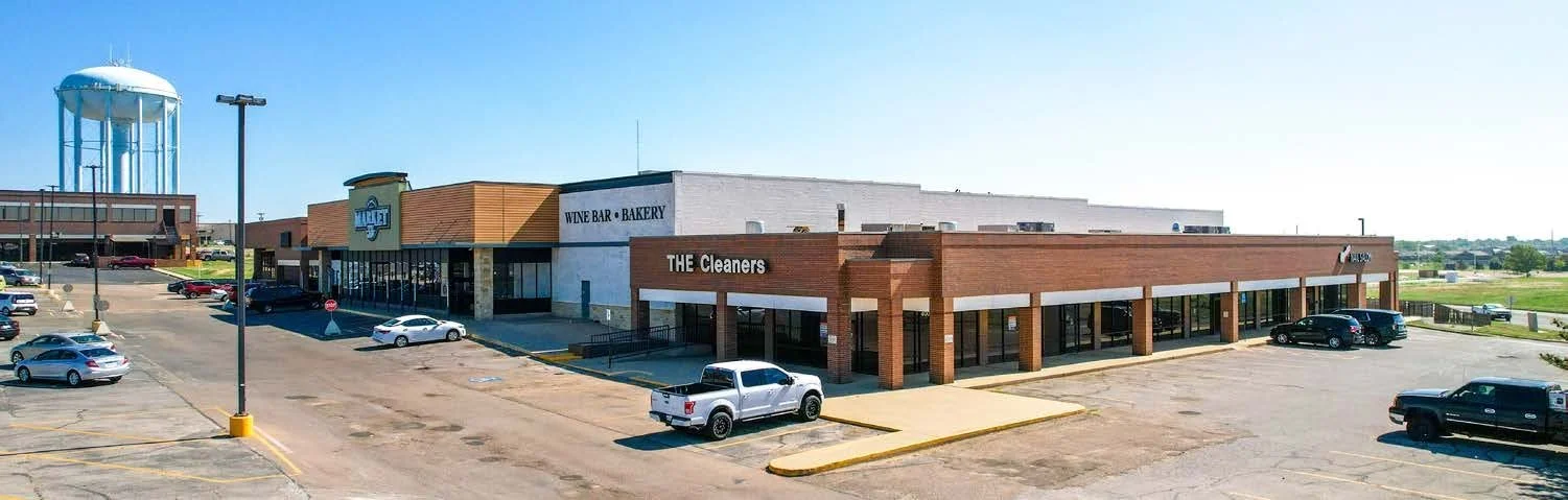 Image of a shopping plaza with a split-level design, featuring stores like The Cleaners, a wine bar, a bakery, and other businesses, with parking spaces in the foreground and a water tower in the background under a clear blue sky.