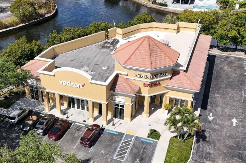A shopping center with Verizon store, Exotic Eyewear, and Optical signs, parking lot with several cars, surrounded by green trees, near a waterway.