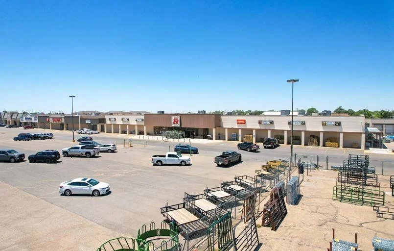 Empty shopping center parking lot with a few cars and shopping carts stacked near the store entrance, under a clear blue sky.