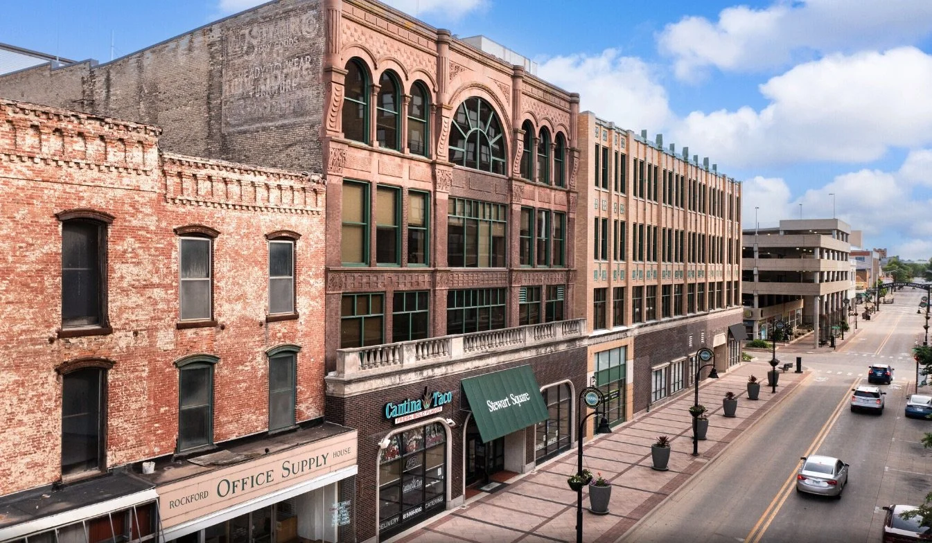 Street view of a downtown area with historic brick buildings, retail stores, and a parking garage under a partly cloudy sky.