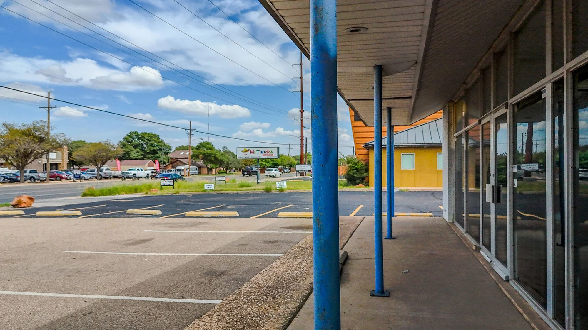 Empty strip mall parking lot with a few parked cars in the background, blue support poles along the sidewalk, and a storefront with large glass windows. A billboard for Mi Tierra is visible in the distance under a partly cloudy sky.