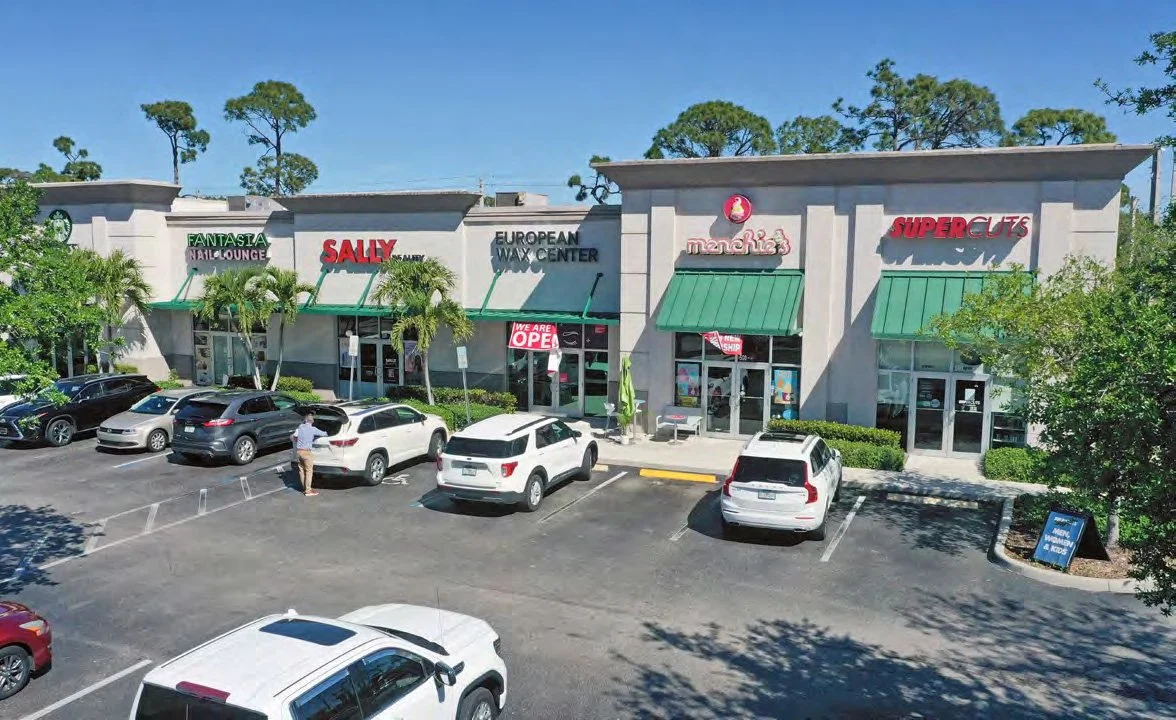 View of a shopping plaza with stores including Sally Beauty, European Wax Center, Menchie's Frozen Yogurt, Super Cuts, and Fantasia All-Lounge, with parked cars in front and a person walking.