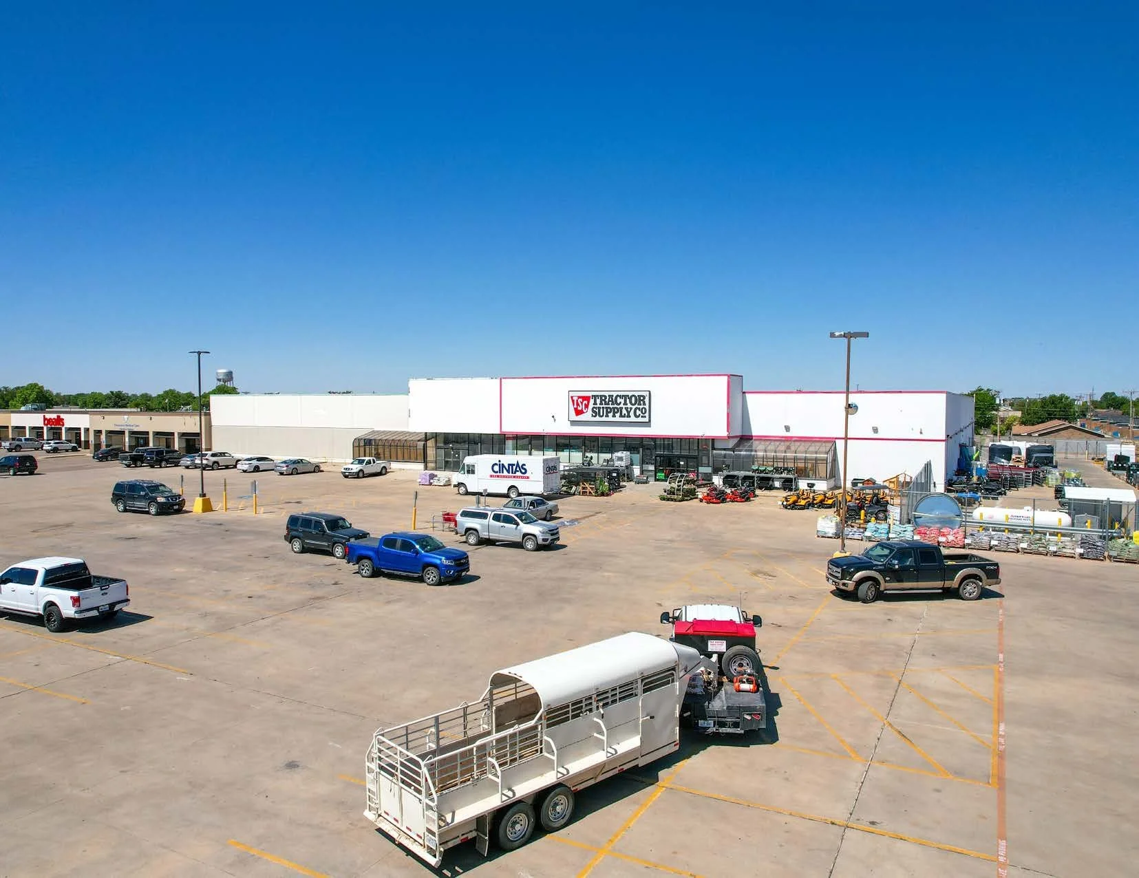 A large parking lot in front of Tractor Supply Co. store, with scattered cars, trucks, and a horse trailers. The store has a white facade with red trim, and large parking spaces marked with yellow lines. Clear blue sky overhead.
