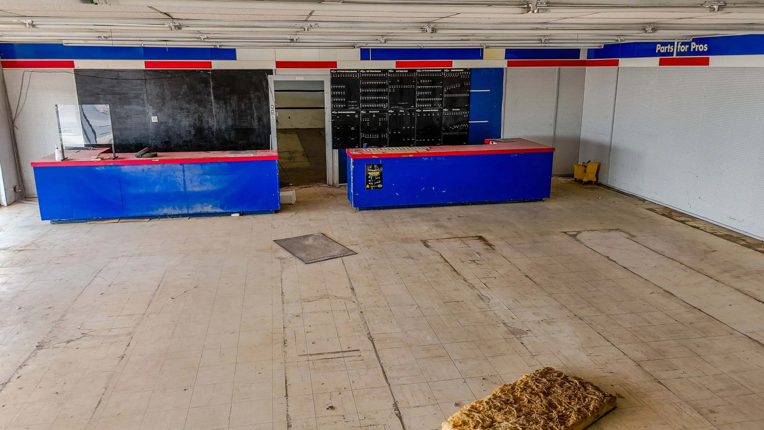 Empty store or workshop space with blue and red counters, a pegboard, a yellow cart, and a worn-out foam mattress on a tiled floor.